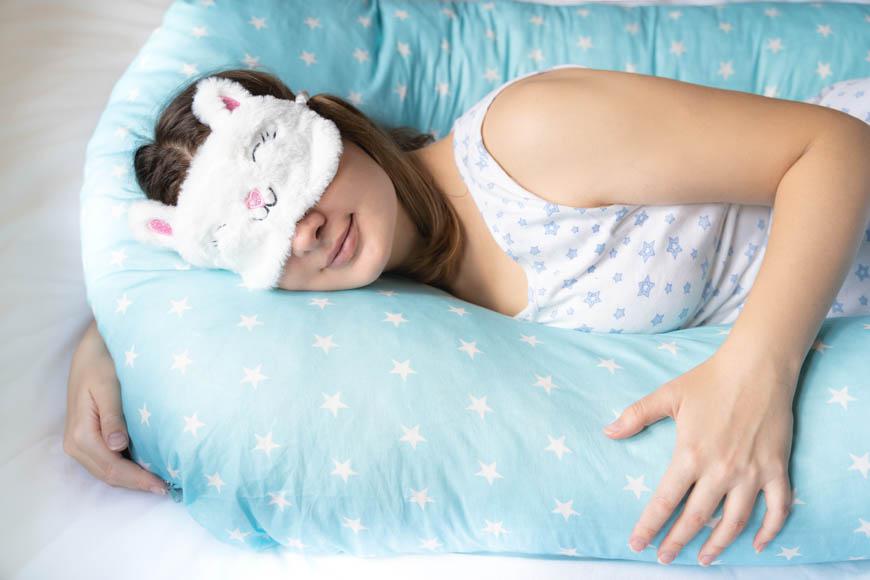 Χαμογελαστή γυναίκα με μάσκα ύπνου κοιμάται σε μαξιλάρι εγκυμοσύνης. Πρακτικό δώρο για μέλλουσα μαμά.