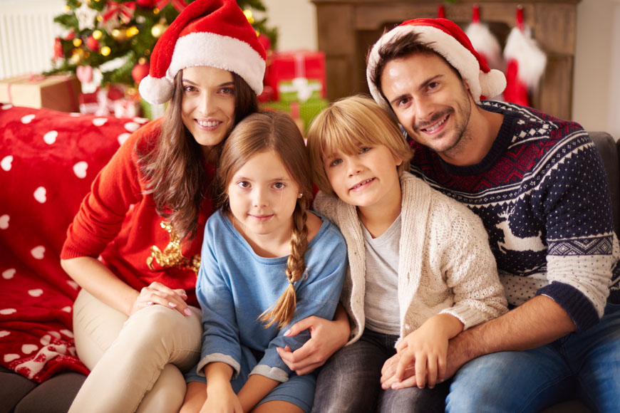 Τετραμελής οικογένεια με Χριστουγεννιάτικα σκουφάκια κάθεται αγκαλιασμένη και χαμογελαστή.